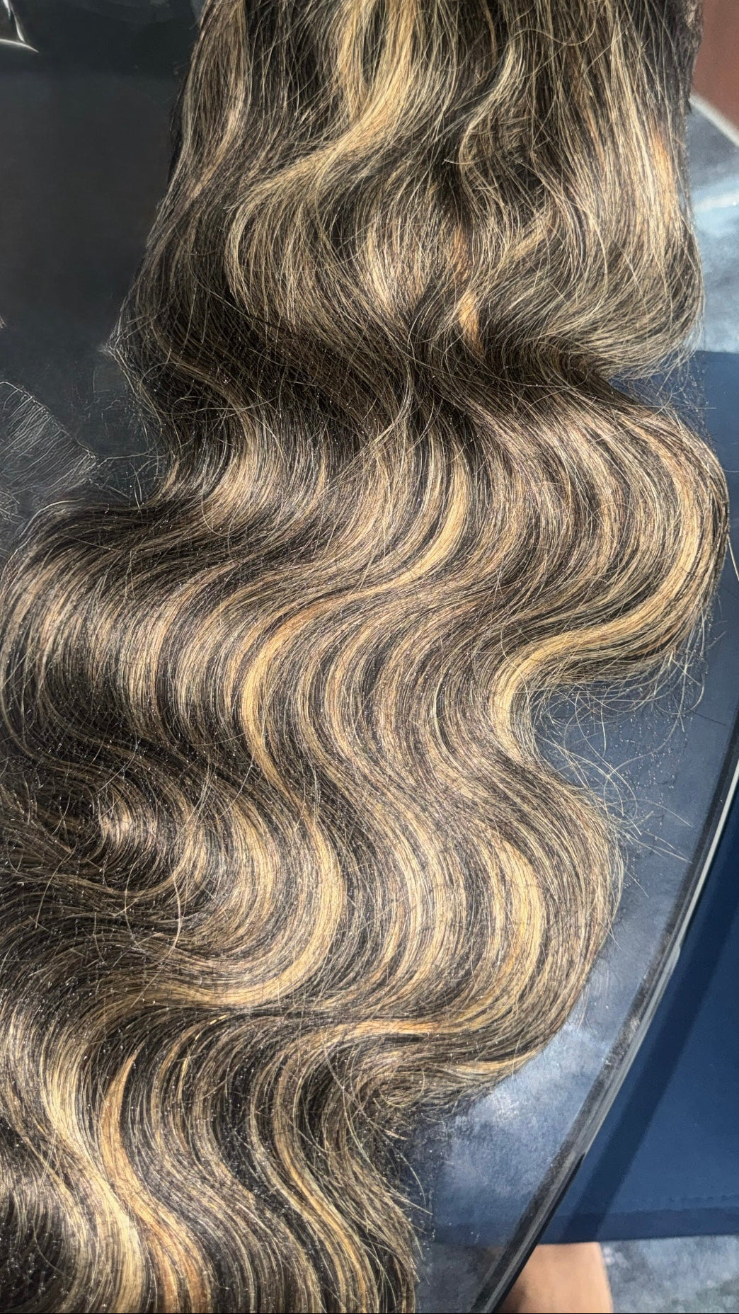 Long, wavy hair extension or wig with a dark brown base and warm blonde highlights, shown draped over a glass surface.
