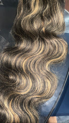 Long, wavy hair extension or wig with a dark brown base and warm blonde highlights, shown draped over a glass surface.